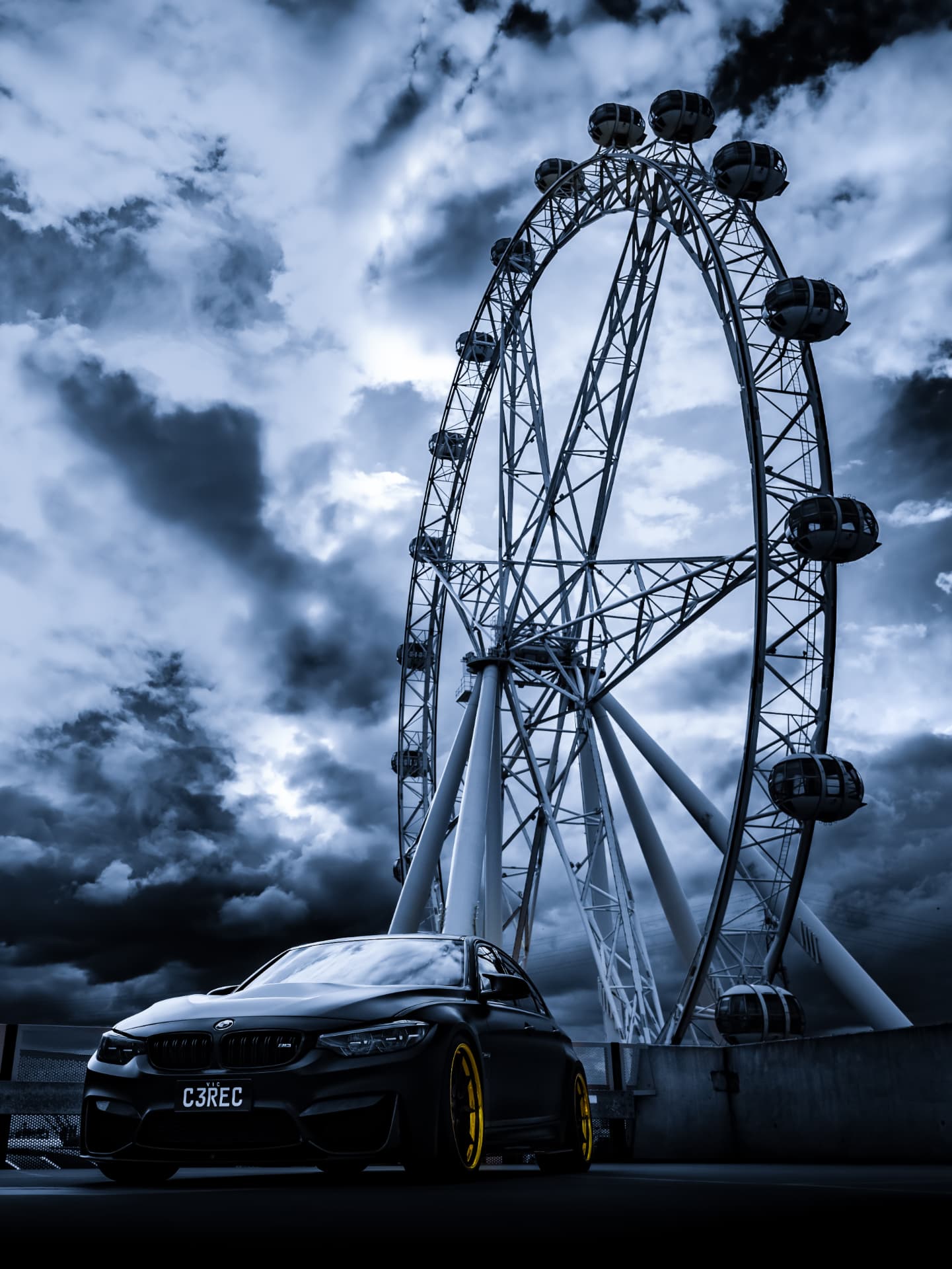 Black BMW M3 parked beneath a ferris wheel under storm clouds
