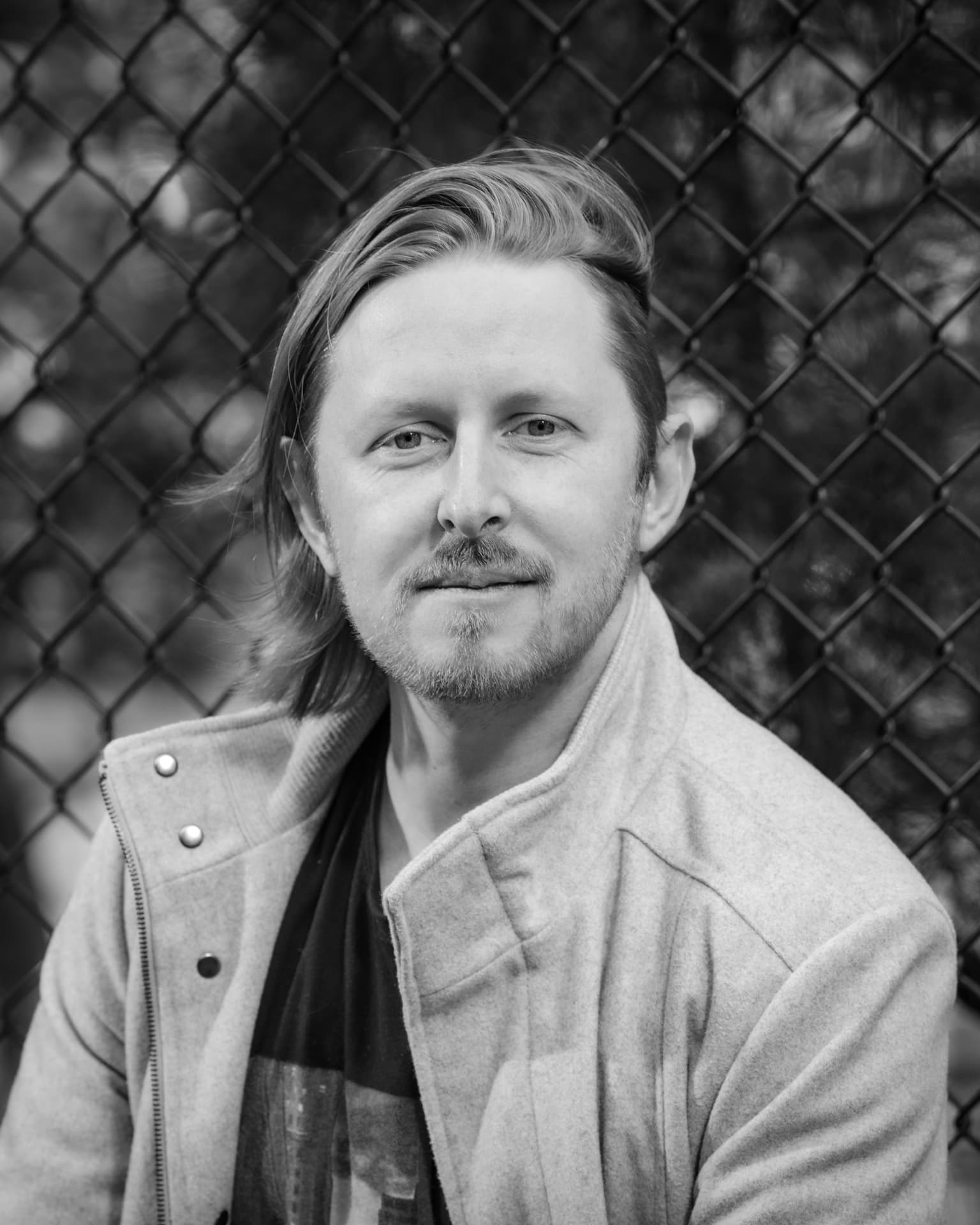 Black and white portrait of a man in a light jacket against a chain-link fence