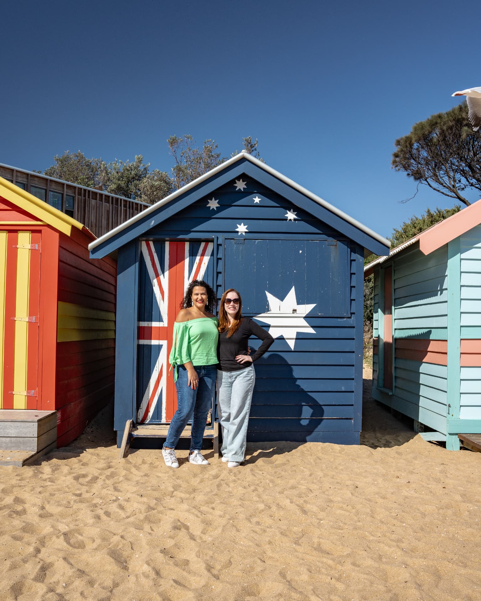 Two friends photographed in front of a Brighton Bathing Box painted with the Australian flag
