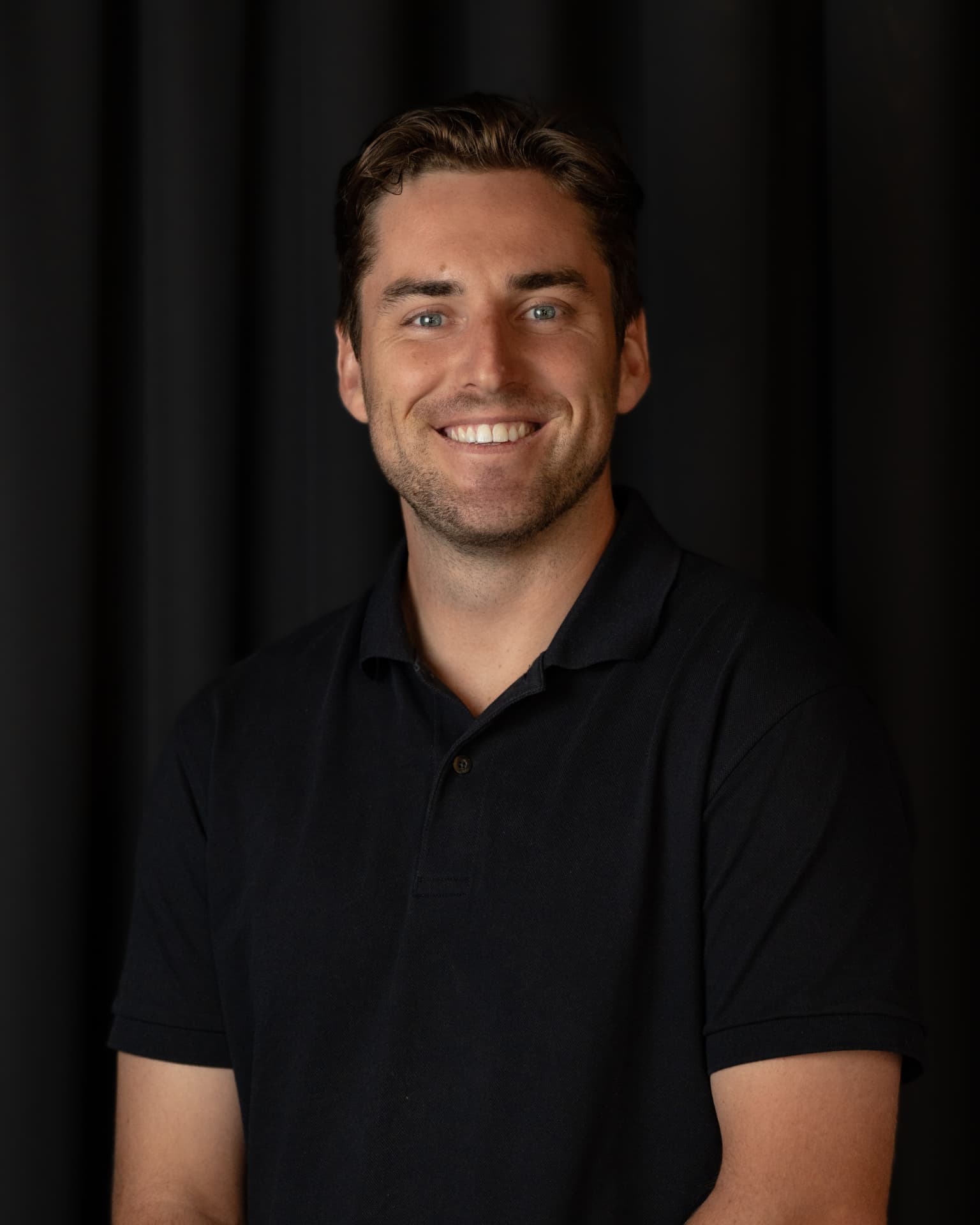 Studio headshot of a smiling man in a black polo shirt against a dark background